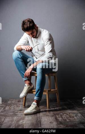 Sad upset young man sitting and looking down over grey background Stock Photo