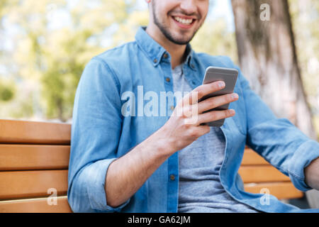 Cropped image of a smiling young man with smartphone sitting on the bench outdoors Stock Photo