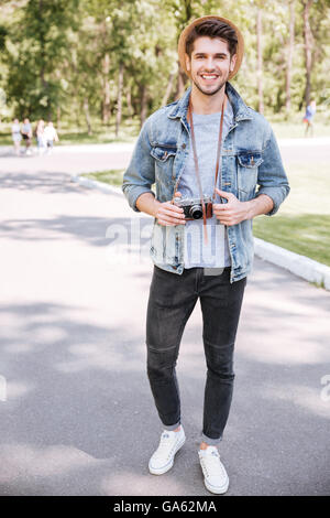 Handsome bearded smiling hipster guy holding retro camera wearing hat in the park outdoors Stock Photo