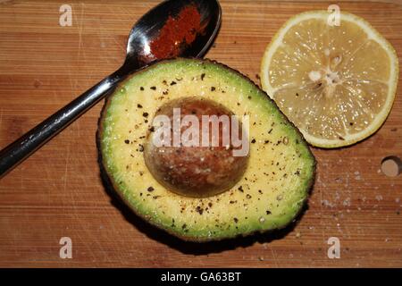 Avocado, stone in, sprinkled with salt and pepper, slice of lemon and spoonful of cayenne pepper at side on wooden board. Stock Photo