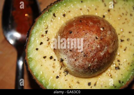 An avocado, stone in, sprinkled with salt and pepper and spoonful of cayenne pepper laid next to it, on wooden board. Stock Photo