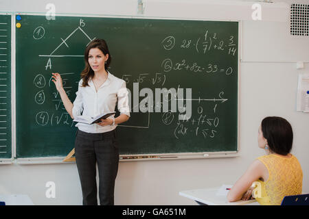 Teacher or docent or educator giving while lesson in front of a blackboard or board a sheet of paper and educate or teaching students or pupils or mates in a school or class Stock Photo