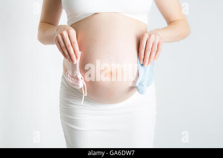 A bregnant woman holds one pink bootie and one blue bootie in front of her baby bump. Stock Photo