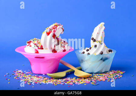 two frozen yogurt with black cherry topping and rainbow sprinkles and chocolate candy on blue background with spoons and sprinkl Stock Photo