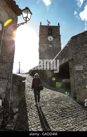 A woman walking down a cobbled street in an ancient village in Europe with a church clocktower behind, against the light with sun flare, high contrast Stock Photo