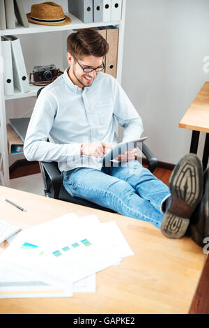 Smiling young man with legs on the table using tablet in office Stock Photo