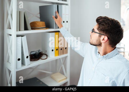 Handsome young businessman in glasses taking folder from shelf in office Stock Photo