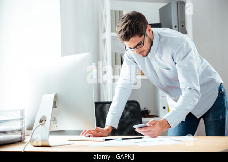 Serious young businessman working with computer and using cell phone in office Stock Photo