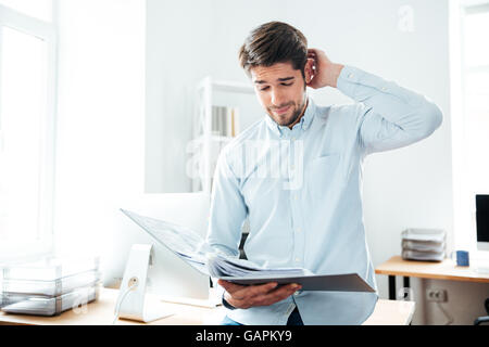 Thoughtful young businessman standing and looking through documents in office Stock Photo
