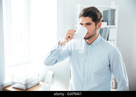 Handsome young man standing and drinking coffee in office Stock Photo