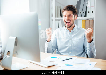Happy excited young businessman sitting on workplace and celebrating success Stock Photo