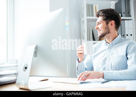 Young smiling handsome businessman drinking coffee and looking away in office Stock Photo