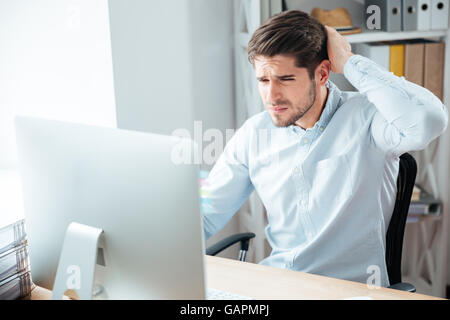 Young thoughtful handsome businessman in blue shirt sitting at the table with laptop in office Stock Photo