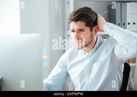 Young thoughtful handsome bearded businessman in blue shirt sitting at the table with laptop in office Stock Photo