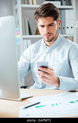 Pensive serious young businessman in casual cloth sitting at the table and using smartphone in office Stock Photo