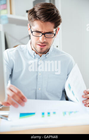 Young pensive handsome businessman sitting on the table and reading document in office Stock Photo