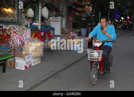 A Chinese man smiles and talks on his smartphone while riding a scooter holding the bar with one hand only, a candy store is on his right. Beijing Stock Photo