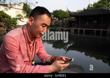 A young Chinese man smiles while playing with his smartphone while sitting by a pond in the Master of Nets Garden. Suzhou, China Stock Photo