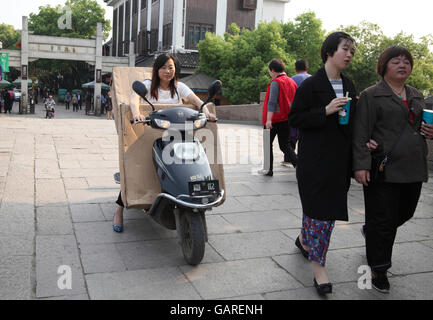 A Chinese woman rides a scooter transporting huge packages she carries in front of her and in the back, other people walk around. Tongli, China. Stock Photo