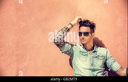 Portrait of handsome man with tattoo on arms sitting and leaning on orange wall in the sun, wearing sunglasses, looking away to Stock Photo
