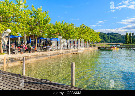 WORTHERSEE LAKE, AUSTRIA - JUN 20, 2015: wooden pier and people sitting at tables along Worthersee lake shore during summer beer Stock Photo