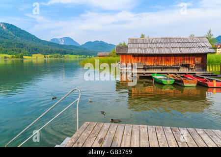 Wooden jetty and fishing boats with wooden houses on shore of Weissensee lake in summer landscape of Carinthia land, Austria Stock Photo