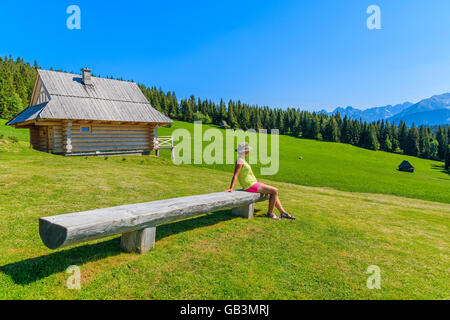 Young woman tourist sitting on wooden bench on green meadow and looking at Tatra Mountains, Poland