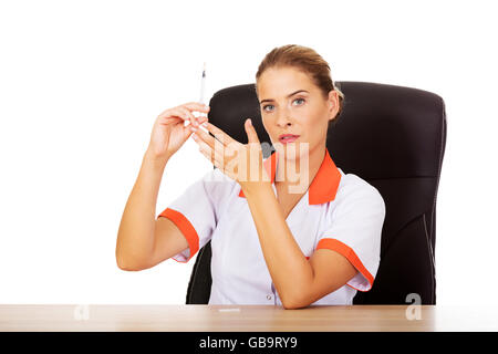 Young female doctor or nurse sitting behind the desk and holding syringe Stock Photo