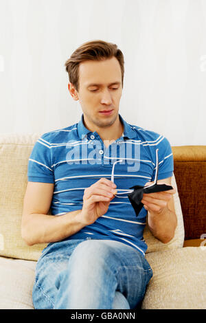 Yong man sitting on couch and cleaning eyeglasses Stock Photo