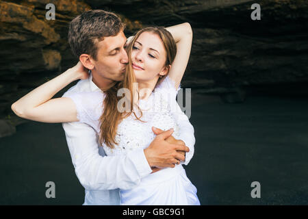 Happy family on honeymoon holiday - just married young man and woman hug with smile on black sand beach with rock background. Ac Stock Photo
