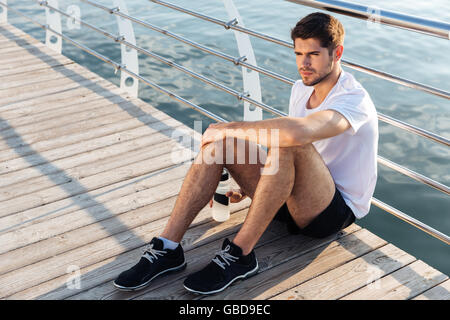 Relaxed young sportsman with bottle of water sitting and relaxing on pier Stock Photo