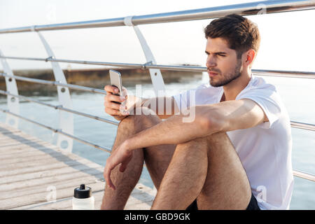 Concentrated young sportsman sitting on pier and using smartphone Stock Photo