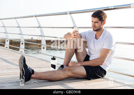 Relaxed young sportsman resting and using mobile phone sitting on wooden terrace Stock Photo