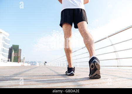 Back view of legs of young man in black shorts and sneakers running on pier Stock Photo