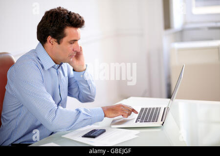 Portrait of a male hispanic boss working on his laptop and speaking on his mobile while sitting on manager desk Stock Photo