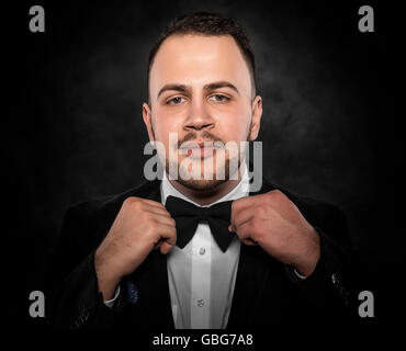 Young handsome man in suit adjusts  bow-tie and smiling on dark background. Stock Photo