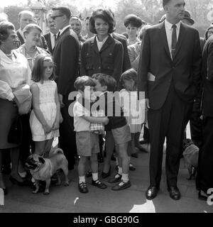 John Kennedy, son of the late American President John F Kennedy, clutches a loaf to his chest and listens as his cousin Anthony Radziwill takes him into his confidence outside the railings of Buckingham Palace forecourt. They were watching the Changing of the Guards before feeding the ducks in St James's Park. Looking on are Jacqueline Kennedy and her daughter Caroline. Stock Photo