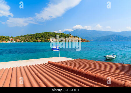 Orange tile roof of a house with Greek flag waving against blue sea background on coast of Samos island, Greece Stock Photo