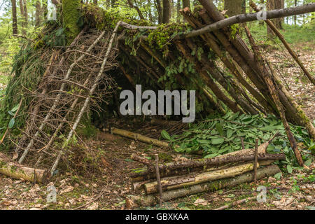 Survival shelter and survivor concept. A-frame shelter construction shelter with log fire reflector in front of shelter opening. Stock Photo