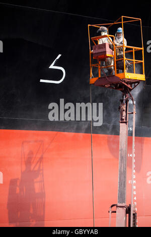 Two suit up pro workers on boom lift is spraying powerful jet of water to clean the vessel hull exterior, their shadow bounce back onto the vessel. Stock Photo