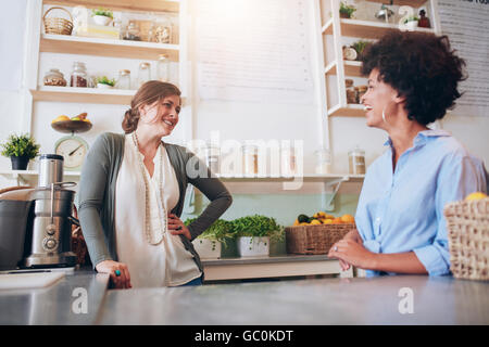 Two young women talking and smiling while working at bar counter. Happy young female employees at fruit juice bar. Stock Photo