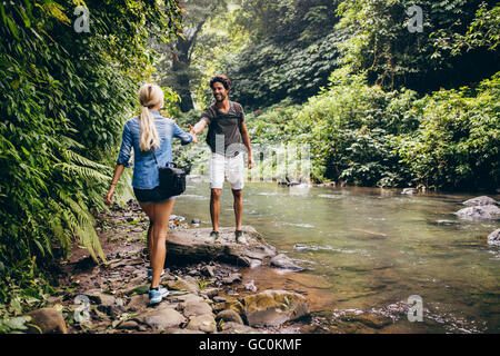 Young couple walking by the mountain stream with man helping his girlfriend. Couple on hikers in forest by the stream. Stock Photo