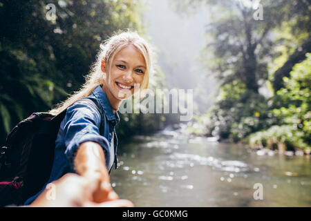 Portrait of happy young woman holding hand of her boyfriend while walking by mountain stream. Couple enjoying a hike in nature. Stock Photo