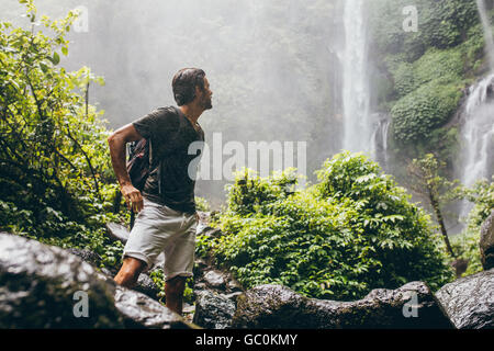 Young man with backpack standing near a waterfall in forest. Male hiker in the nature during rain. Stock Photo