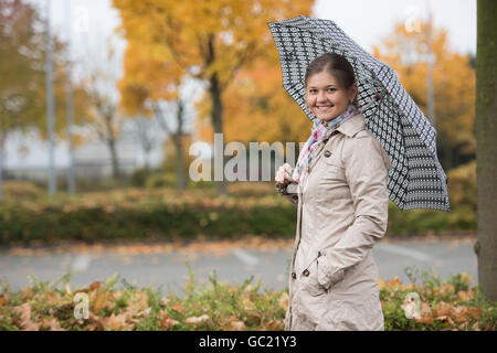 Young woman standing under umbrella in autumn park, wearing beige mackintosh Stock Photo