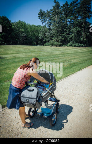 Mother looks into the pram on walk in the park. Rear view. Stock Photo