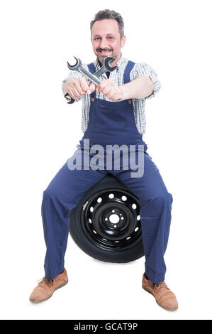 Smiling handsome mechanic with wrench sitting on tire as auto services concept isolated on white background Stock Photo