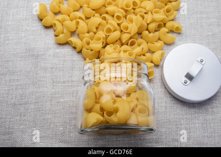 raw pasta spilled out of the glass jar with grey cap, of linen tablecloths Stock Photo