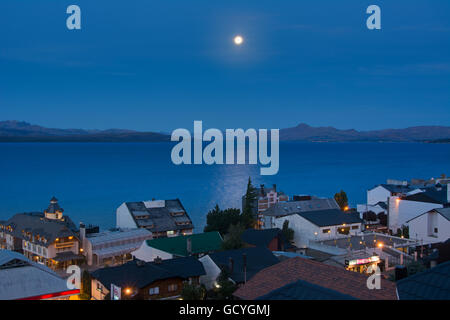 The moon rising over a lake from above the town roof tops showing some yellow city lights against the blue scene; Bariloche, Argentina Stock Photo