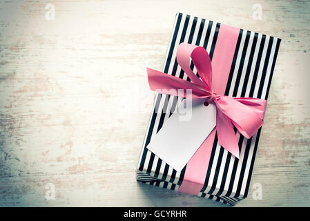 Gift box wrapped in black and white striped paper with pink ribbon on a white wood old background. Empty note tied over. Vintage Stock Photo
